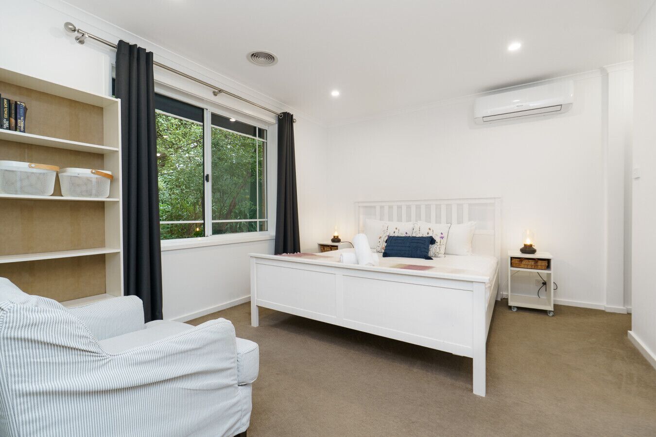 A bedroom with a white bed, a bedside table with a lamp, a window with curtains, a bookshelf, and a white air conditioning unit.