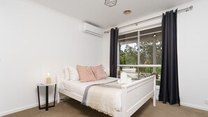 A bedroom with a bed, a bedside table, an air conditioning unit, a window with curtains, and a view of greenery outside.