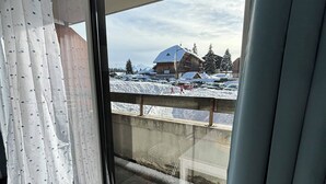 Ett rum med ett stort fönster, ett trägolv och utsikt över en byggnad med ett snötäckt tak.