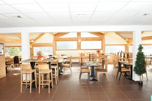 Ein geräumiges Restaurant mit hohen Decken und großen Fenstern, Holztischen und -stühlen sowie einem Blumentopf.