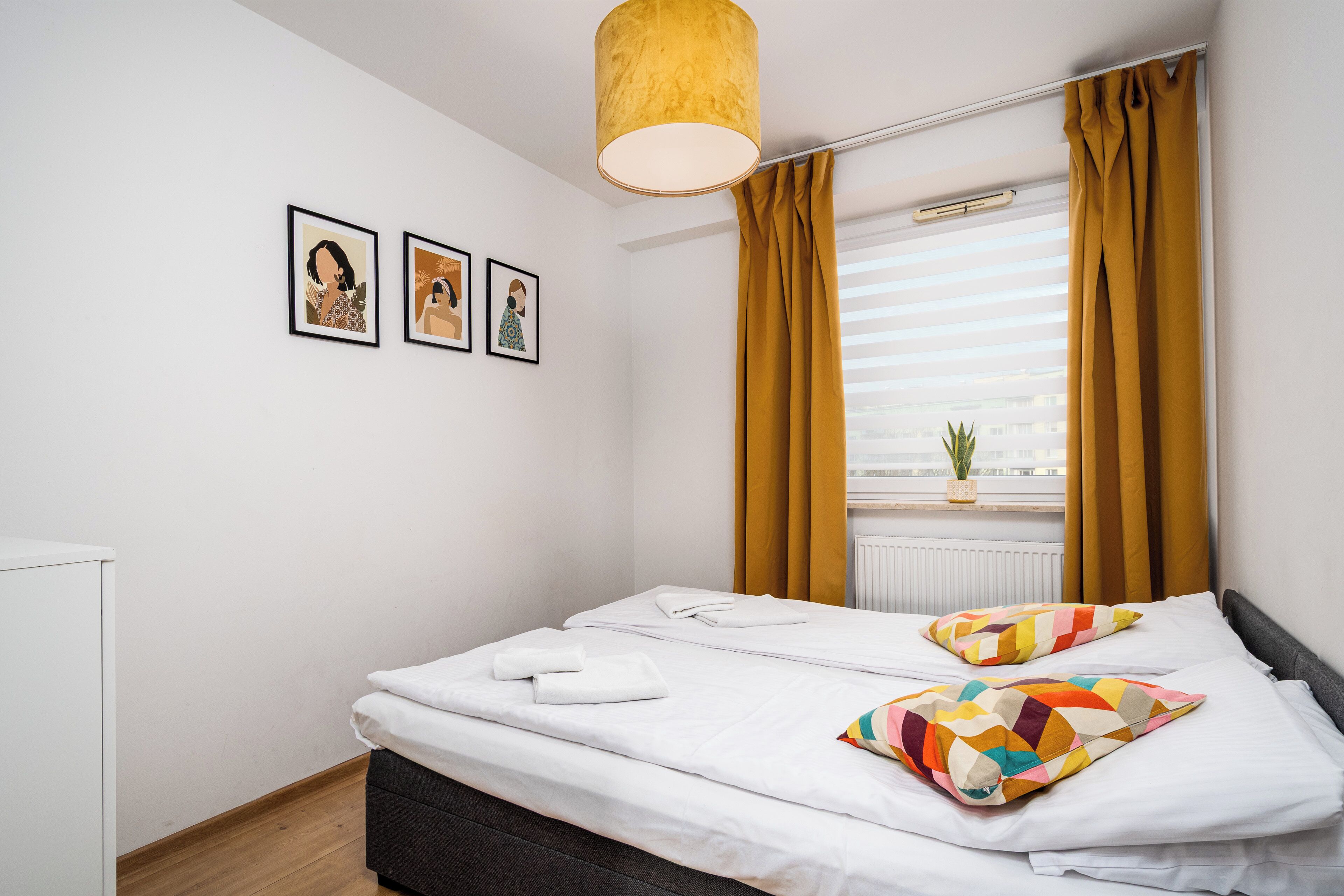A neatly made bed with white linens, a gray headboard, and a colorful pillow. There are three framed pictures on the wall, a window with yellow curtains, and a hanging light fixture.