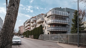 Una calle residencial con edificios de apartamentos modernos, una acera bordeada de árboles y una valla metálica.