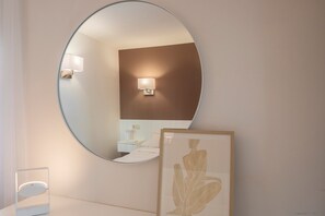 Een ronde spiegel aan de muur, een bedlamp, een kunstwerk in een lijst en een nachtkastje met een lamp.