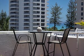 A balcony with two chairs and a table, overlooking a high-rise building and a body of water. - Rainbow Sky Terrace - Ocean Views & Private Spa (Coolangatta)
