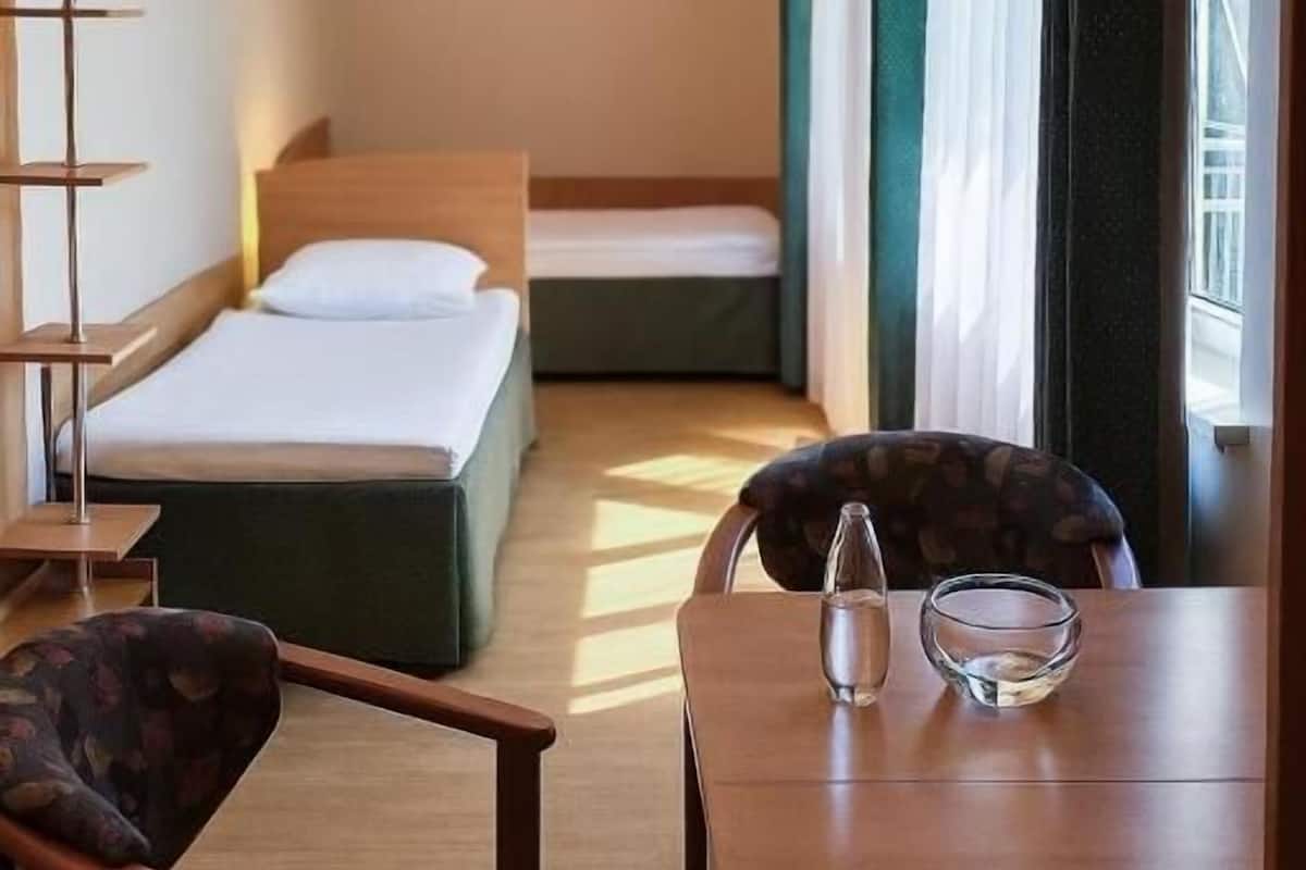 Ein Hotelzimmer mit einem Bett, einem Tisch mit Stuhl, einem kleinen Tisch mit Wasserflasche und Glas sowie einem Fenster mit Vorhängen.