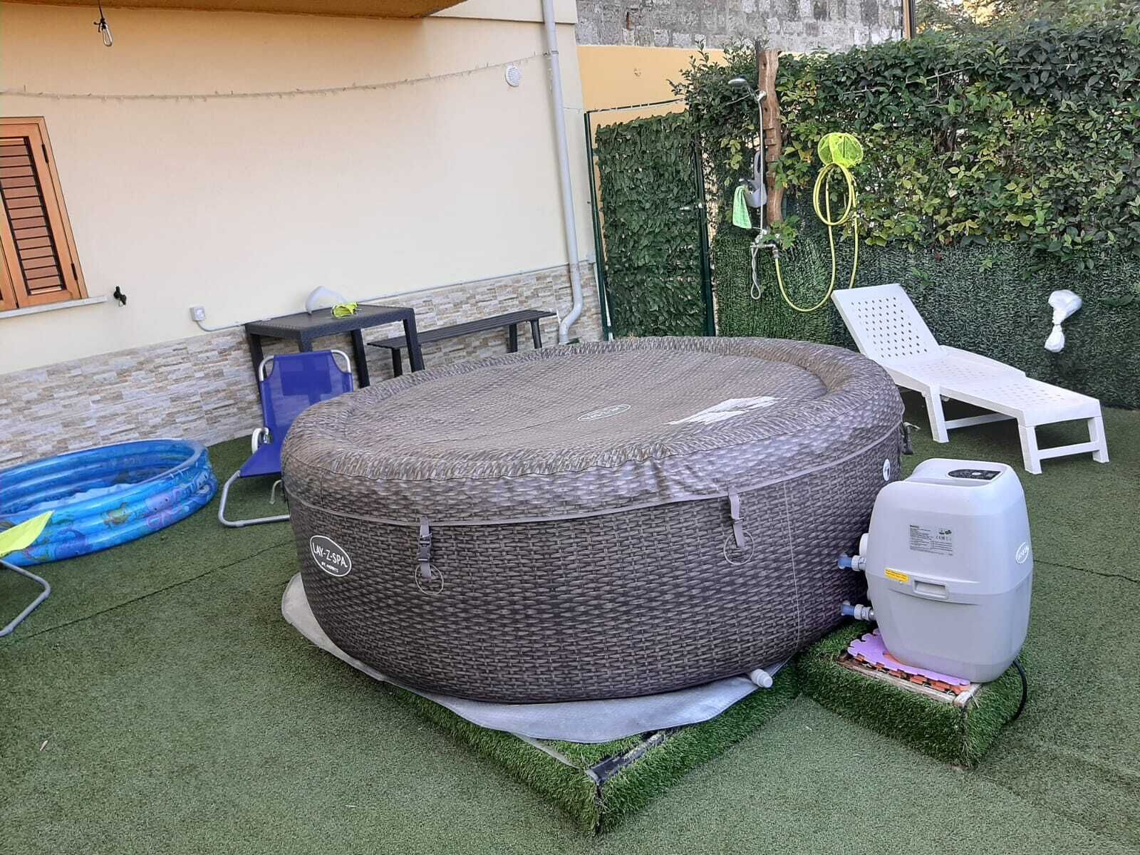 A hot tub with a wicker cover, a blue inflatable pool, and a grill on artificial grass.