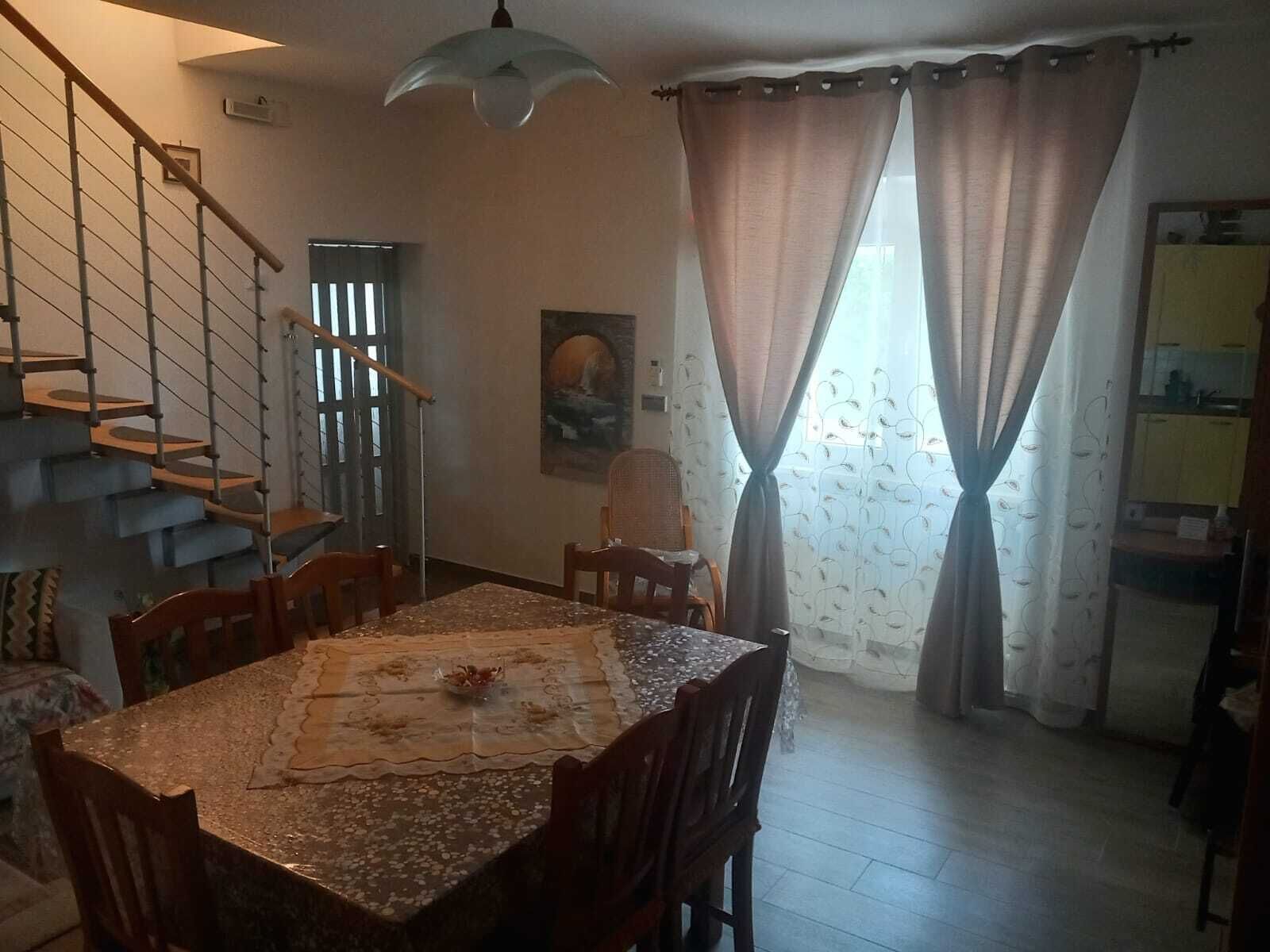 Een eetgedeelte met een tafel, stoelen, een schilderij aan de muur, een trap en een raam met gordijnen.