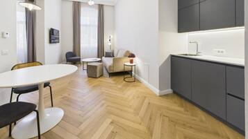 Een moderne hotelkamer met een visgraat parketvloer, een ronde eettafel, een bank, een televisie en een kitchenette.