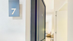 Un pasillo con una puerta numerada con el número 7, que da a una habitación con una cama y una silla.