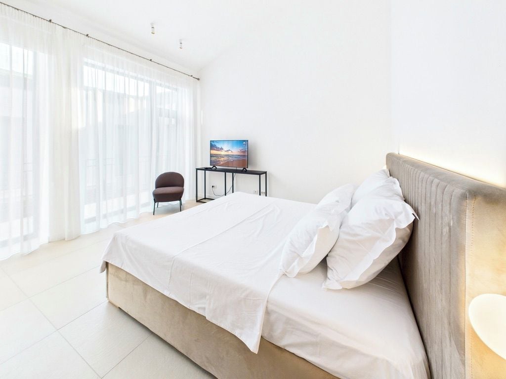 Habitación de hotel con una cama grande, una silla, una mesita con un cuadro y una ventana con cortinas translúcidas.