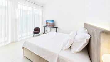 Habitación de hotel con una cama grande, una silla, una mesita con un cuadro y una ventana con cortinas translúcidas.