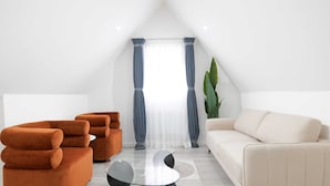 A modern living room with a white ceiling, a large window with curtains, a beige sofa, and orange armchairs. - The Wembley Crown Heights 1 Bed Flat  (Wembley)
