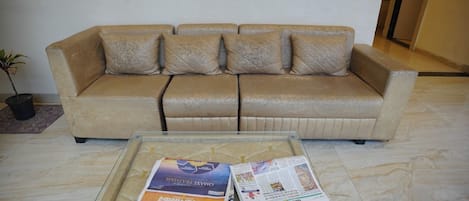 A beige sofa with four cushions, a glass coffee table with magazines, and a potted plant to the left.