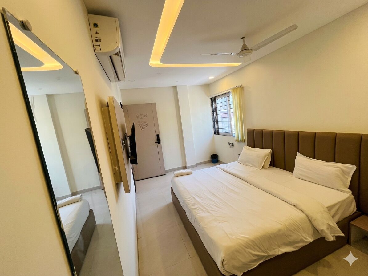 A hotel room with a large bed, a ceiling fan, an air conditioning unit, a window with curtains, and a door leading to another room.
