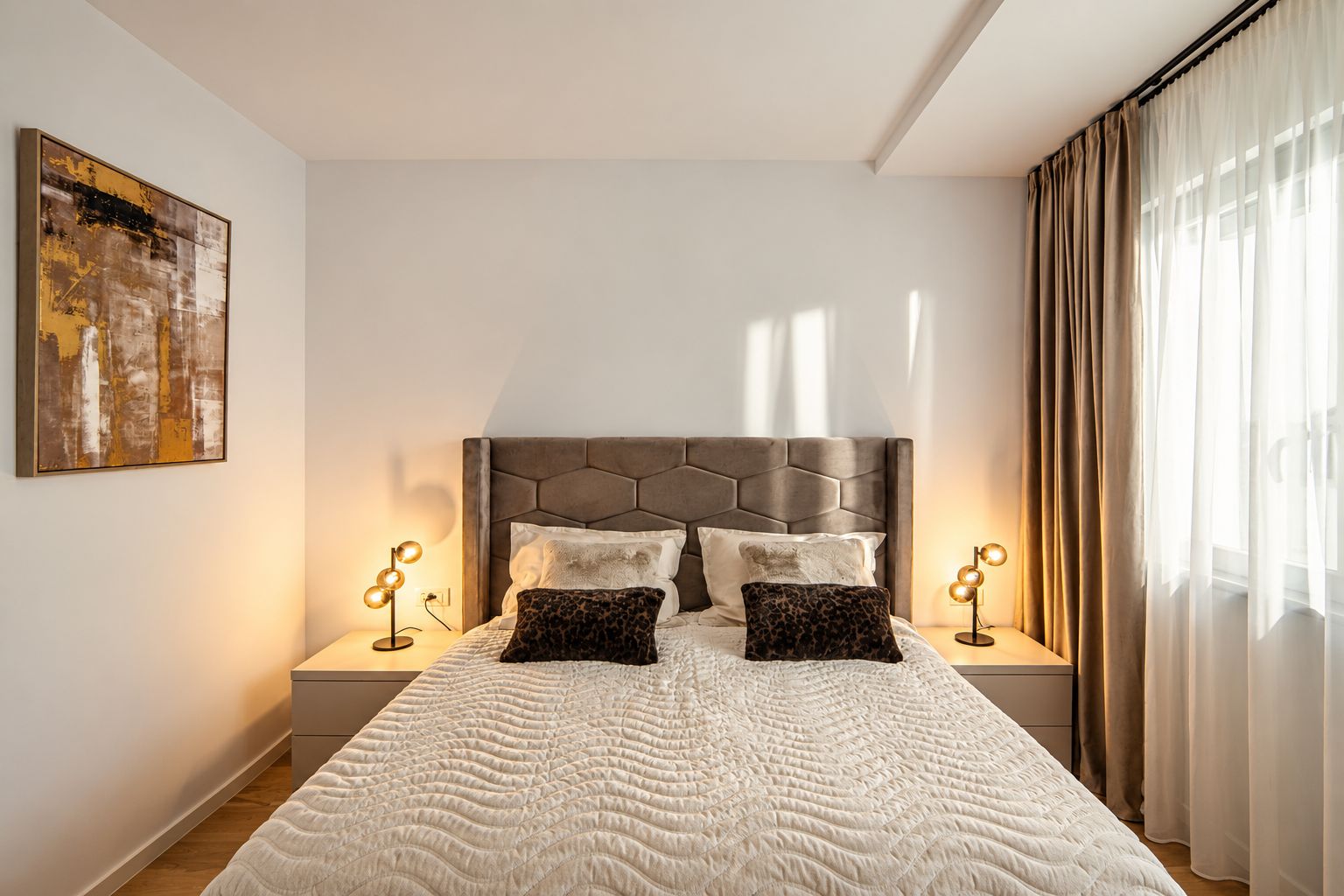 A neatly made bed with a textured white duvet, a geometric headboard, and two bedside lamps with round lights.
