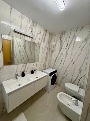 Une salle de bain moderne avec des murs en marbre, un meuble-lavabo à deux vasques, un lave-linge et des toilettes.