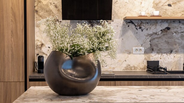 A modern kitchen with a marble countertop, a large vase with flowers, and a wooden cabinet. - MaisonA (Timișoara)