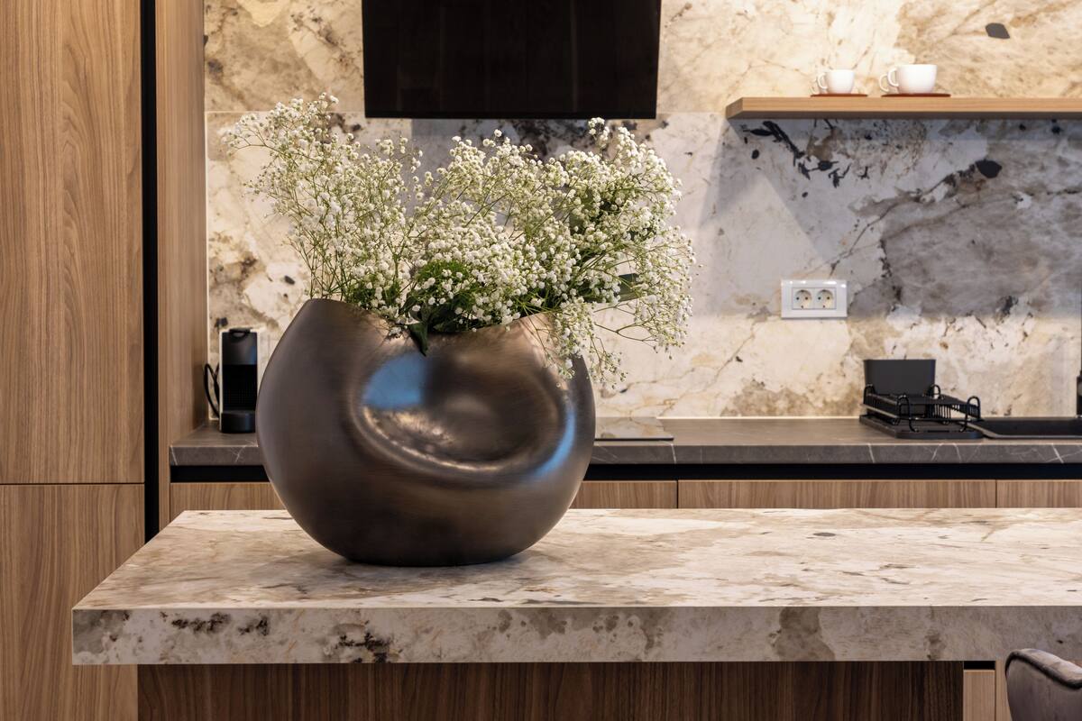 A modern kitchen with a marble countertop, a large vase with flowers, and a wooden cabinet.