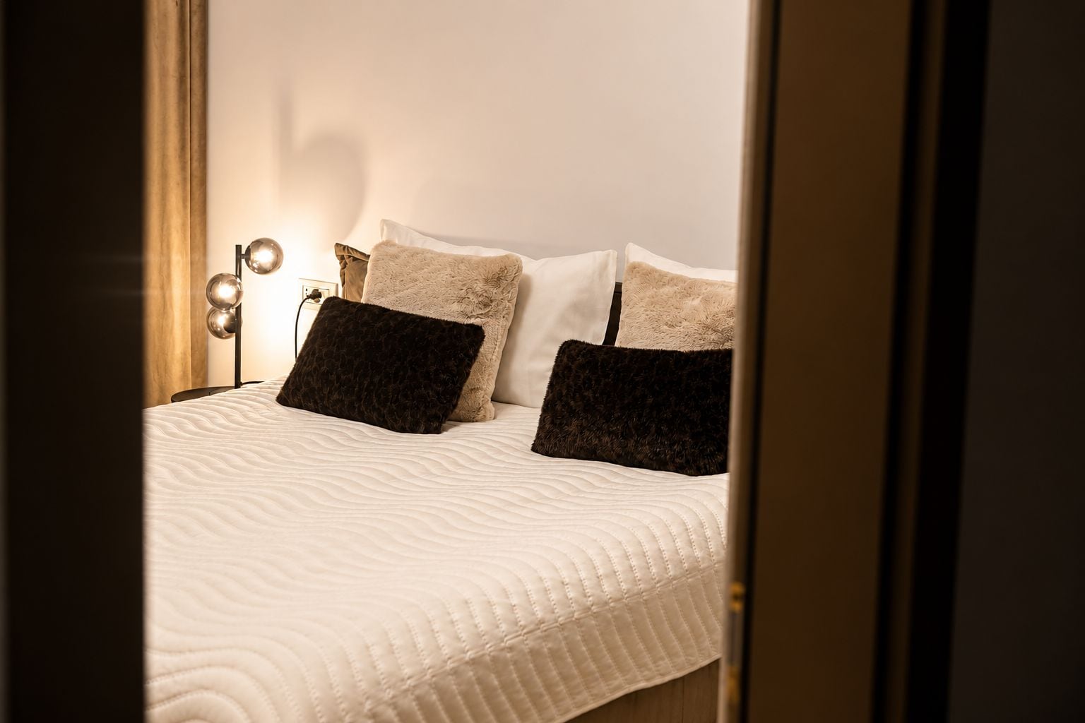 Una cama bien hecha con colcha blanca, dos almohadas oscuras y una lámpara de noche con varias bombillas.