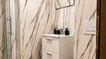 Un baño con pared de mármol, cabina de ducha de vidrio, lavamanos blanco y un inodoro.