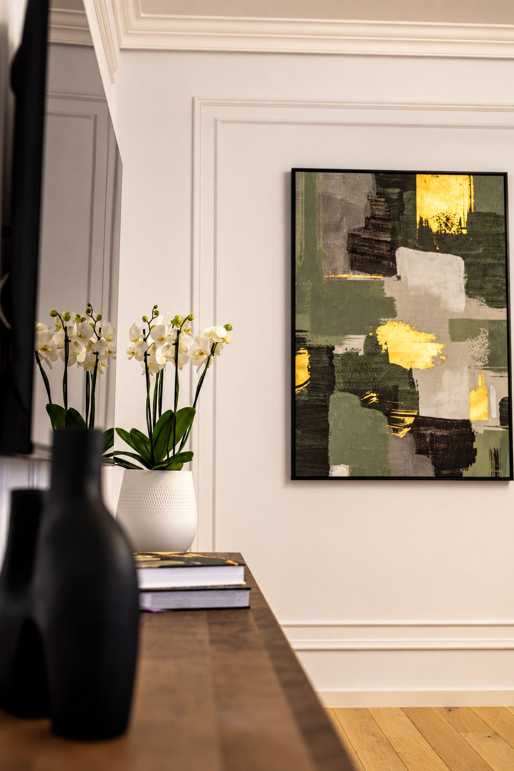 Una habitación con una mesa de madera, un jarrón blanco con flores blancas y un cuadro abstracto enmarcado en la pared.