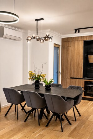 A modern dining area with a round table, six grey chairs, a black vase with flowers, and a pendant light fixture. - MaisonA (Timișoara)