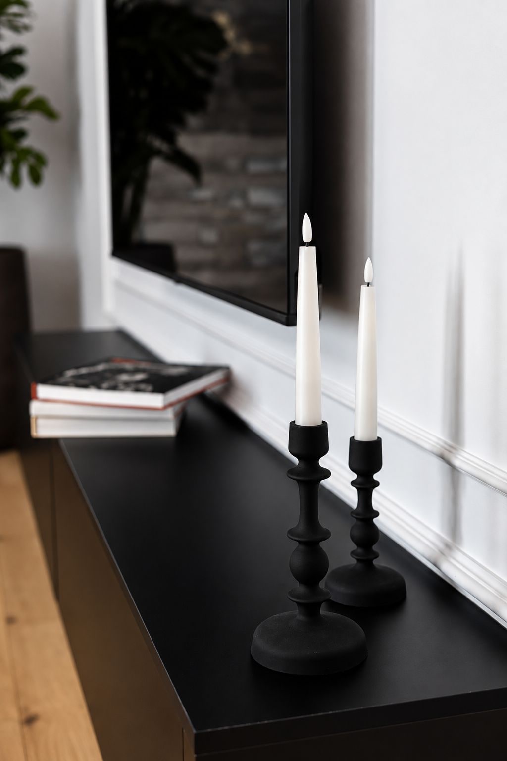 A black table with two white candles on black candle holders, a book, and a television in the background.