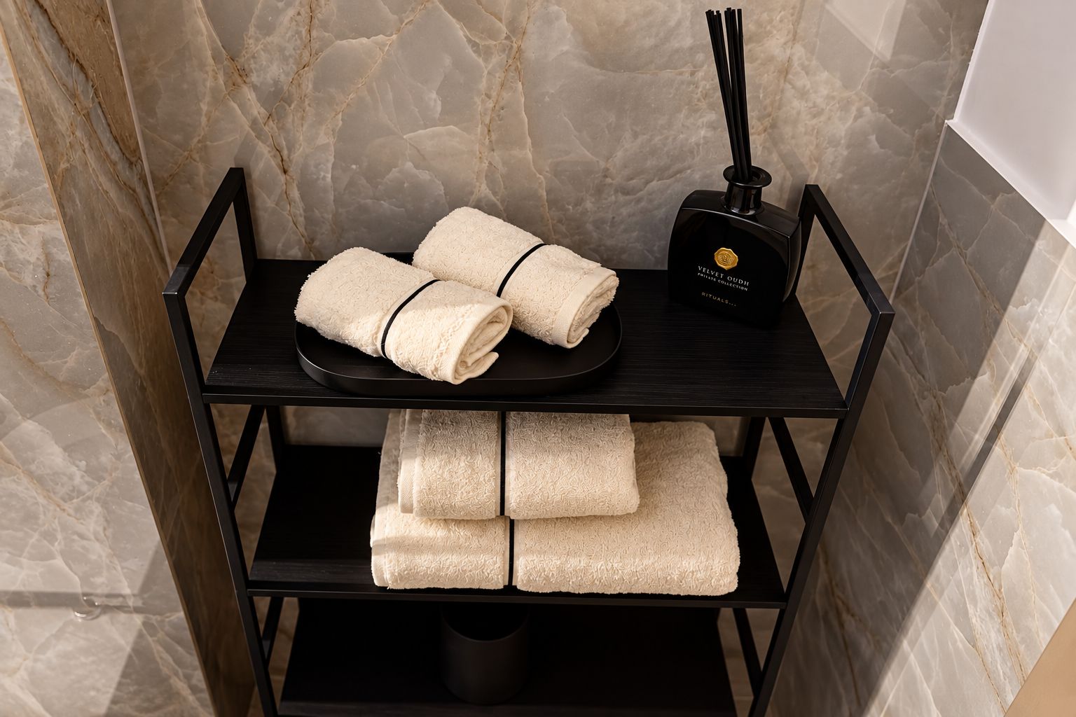 A black metal shelf with two folded towels and a black box with a reed diffuser in a bathroom with marble walls.