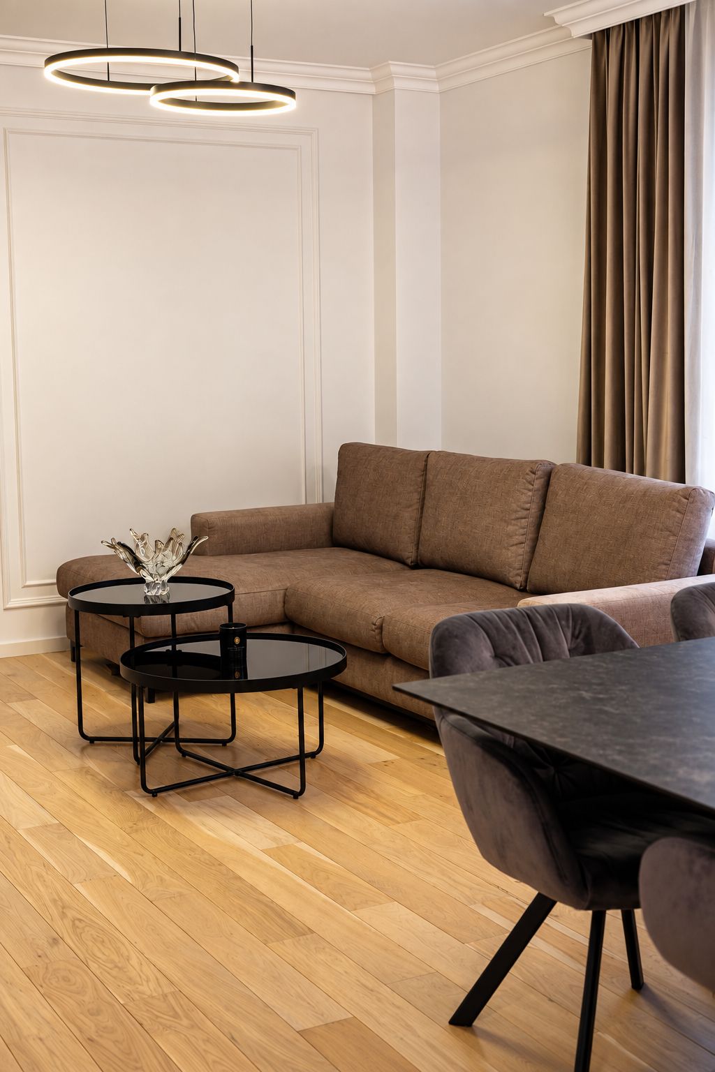 A modern living room with a brown sectional sofa, two black round tables, and a dining table with chairs.