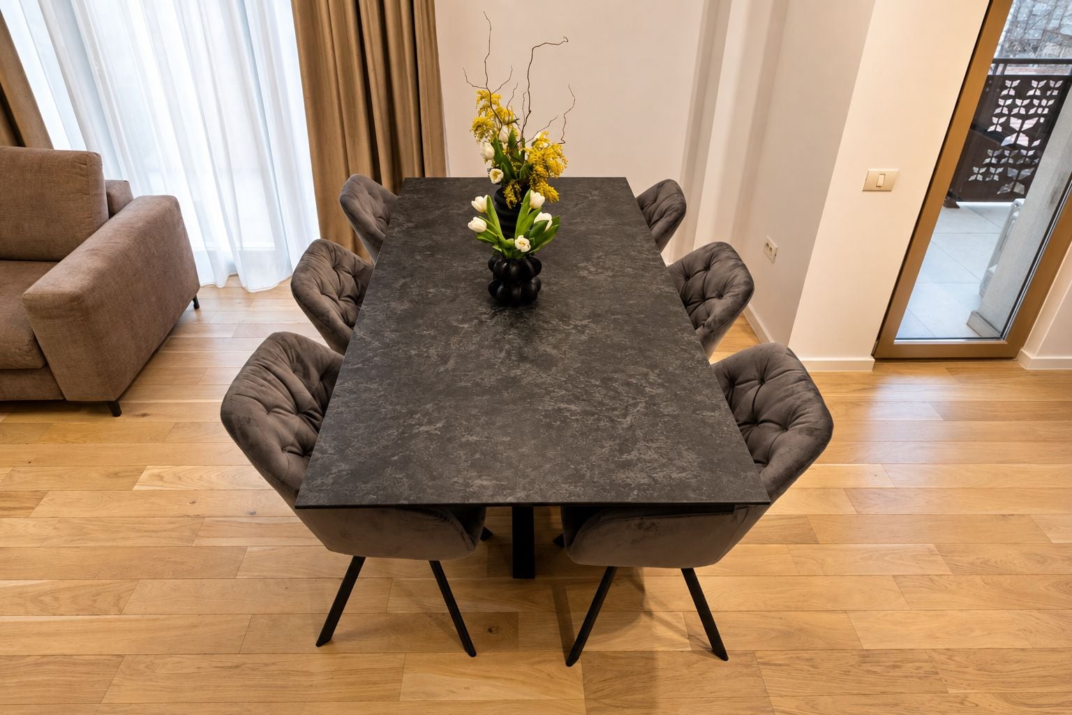 A dining table with a dark surface and six upholstered chairs, a vase with flowers, and a wooden floor.