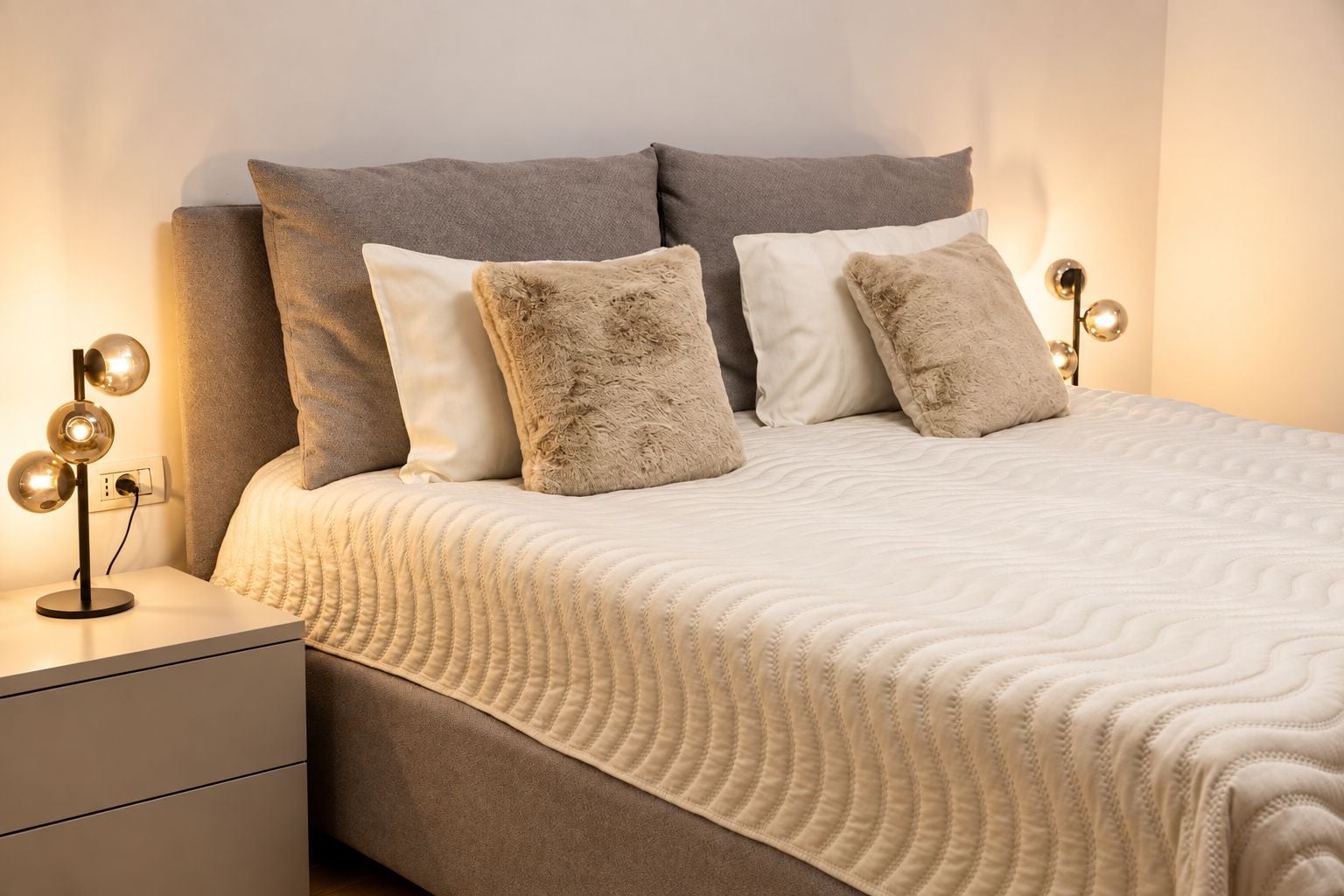 Un lit bien fait, recouvert d’une couette blanche à texture intéressante, flanqué de deux tables de chevet avec des lampes, et doté d’une tête de lit grise.