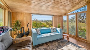 A room with a wooden ceiling, a blue sofa, a wicker chair, and a view of greenery outside. - 864 Sabal Court - Beautiful Marsh Views (Fripp Island)
