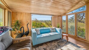 A room with a wooden ceiling, a blue sofa, a wicker chair, and a view of greenery outside. - 864 Sabal Court - Beautiful Marsh Views (Fripp Island)
