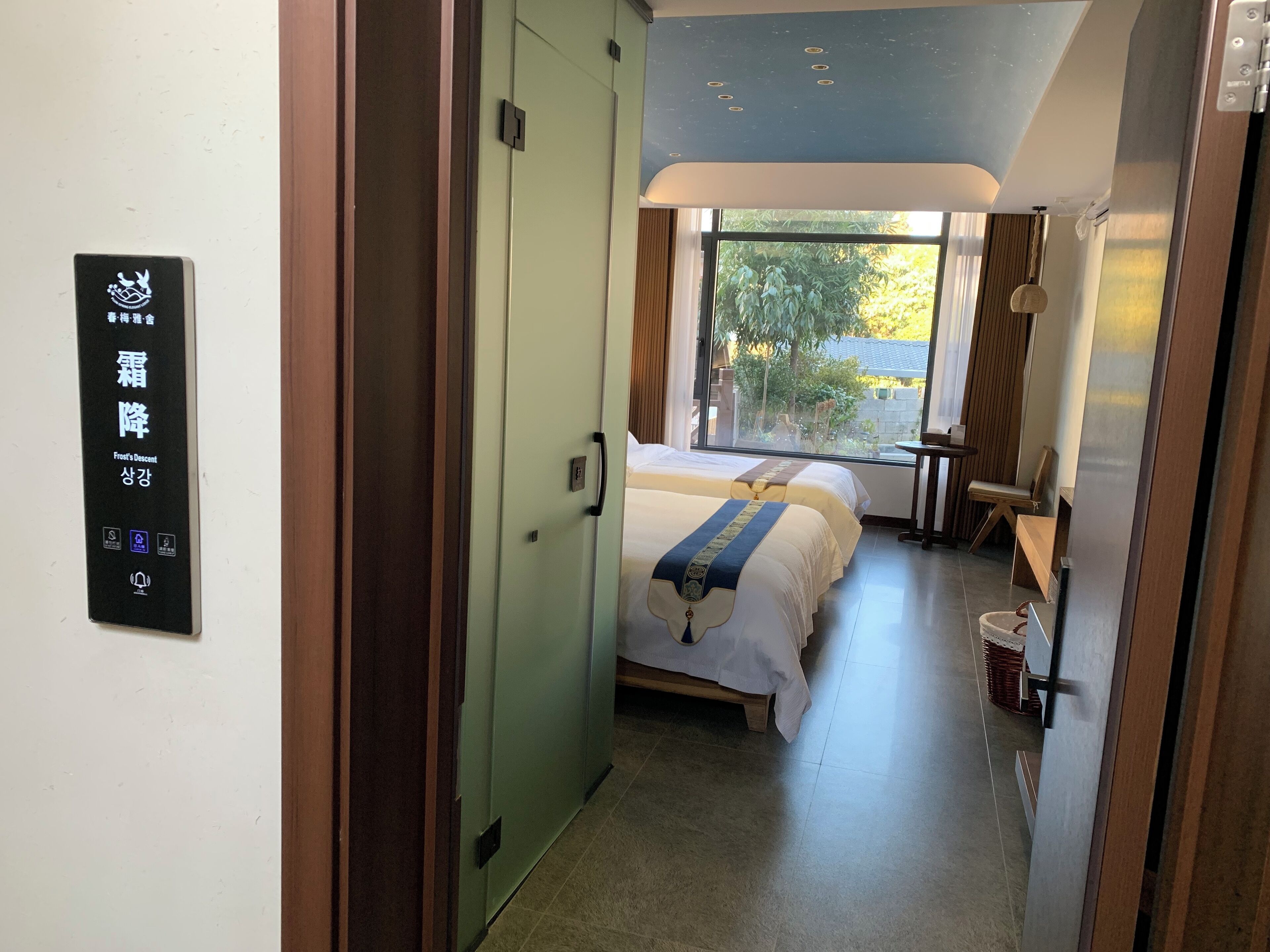Ein Hotelzimmer mit Bett, Fenster mit Aussicht, einer Tür mit Schild und einem kleinen Tisch mit Stuhl.
