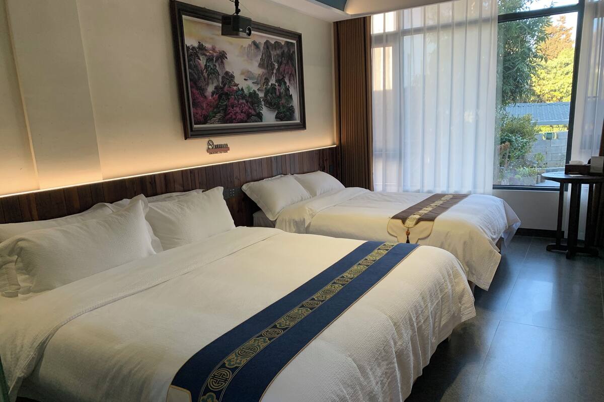 A hotel room with two beds, a large window with curtains, a painting on the wall, and a small table by the window.
