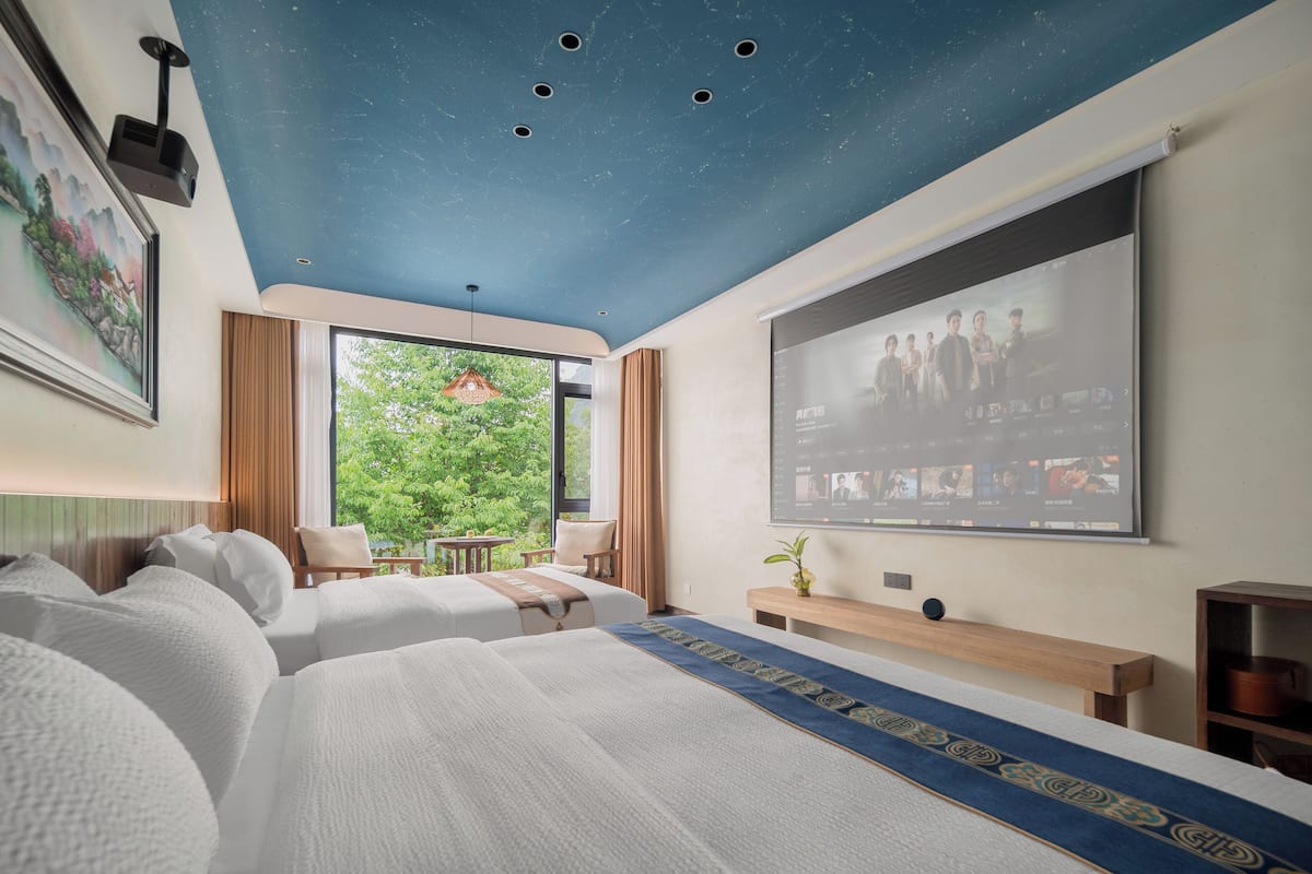 A hotel room with two beds, a ceiling projector, a large window with a view of greenery, and a screen displaying a group of people.