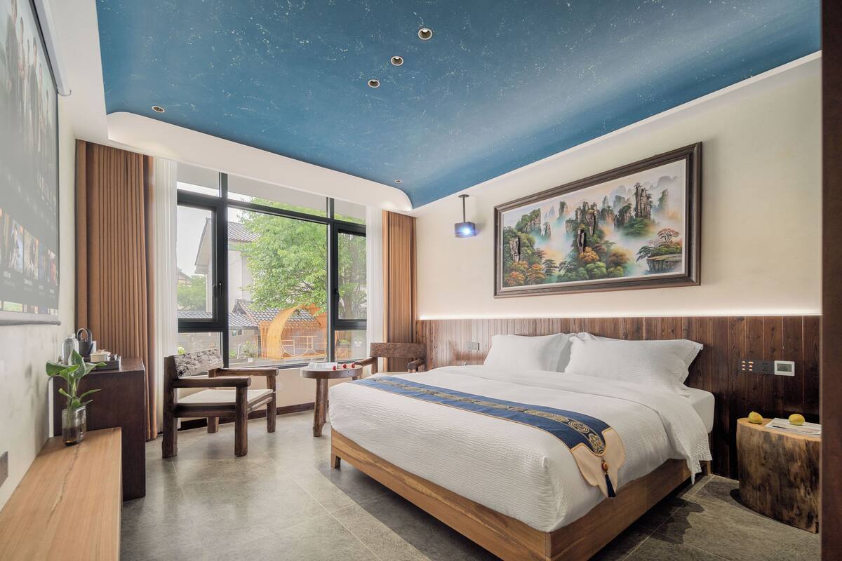 A bedroom with a large bed, a desk, a chair, a window with a view, and a painting on the wall.