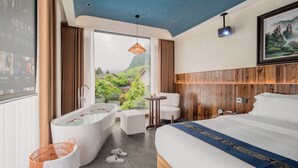 A modern hotel room with a large bed, a bathtub, a small table, and a view of greenery outside. - Zhangjiajie Stone Forest Retreat (Zhangjiajie)