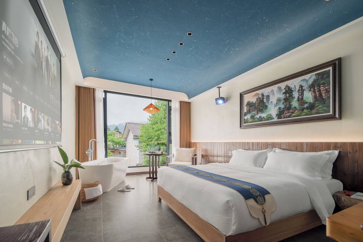 A modern hotel room with a large bed, a bathtub, a desk, and a large window.