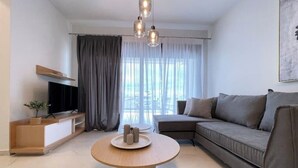 Een moderne woonkamer met een hoekbank, een ronde houten salontafel, een flatscreen televisie op een houten dressoir en dunne gordijnen voor het raam.