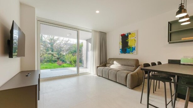 A modern living room with a sofa, a dining table with chairs, a TV mounted on the wall, and a large sliding glass door leading to a garden. - Le Residenze Di Zelo - Private Parking (Mestre)