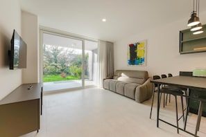 Uma sala de estar moderna com sofá, mesa de jantar com cadeiras, TV fixada na parede e uma grande porta de vidro deslizante que dá para um jardim.