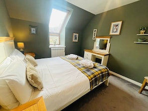 A neatly made bed with pillows and a plaid blanket, a small bedside table with a lamp, a window with a view, and a shelf with decorative items.