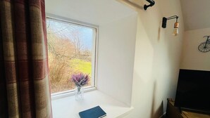 Una habitación con ventana, una pequeña mesa, un jarrón con flores, un libro, una lámpara de pared y una decoración con bicicleta.