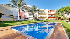 A swimming pool with a tiled deck, surrounded by a well-maintained lawn and palm trees, with multi-story buildings in the background. - Sunny Village Vilamoura (Loulé)