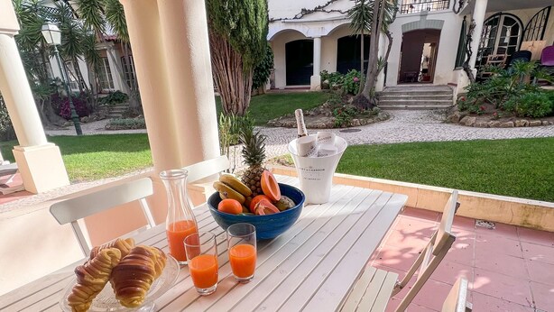 A table with a bowl of fruit, a croissant, and glasses of juice on a patio with a view of a building and garden. - Sunny Village Vilamoura (Loulé)