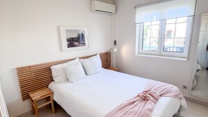 A bedroom with a bed, wooden headboard, bedside table, nightstand, and a window with a view. - Sunny Village Vilamoura (Loulé)