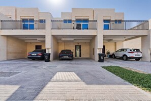 A multi-story residential building with a central entrance, two cars parked in front, and a small garden area. - Elegant 3BR Villa Retreat w Private Backyard (Damac Hills 2)