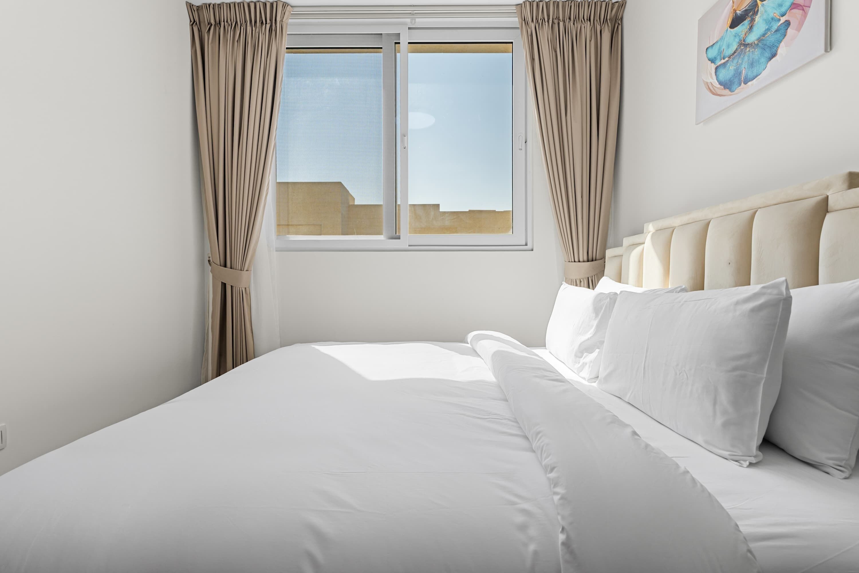 A neatly made bed with white linens, a headboard, and a window with curtains.
