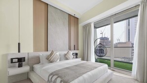 A modern bedroom with a large bed, a nightstand, a wall-mounted lamp, and a view of a construction site through the window. - Stylish 2BR w Study and City Views (Dubai Marina)